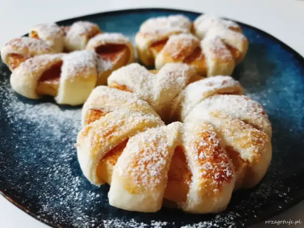 Pyszne ciasto francuskie z jabłkami, posypane cukrem pudrem, prezentuje się apetycznie na granatowym talerzu.