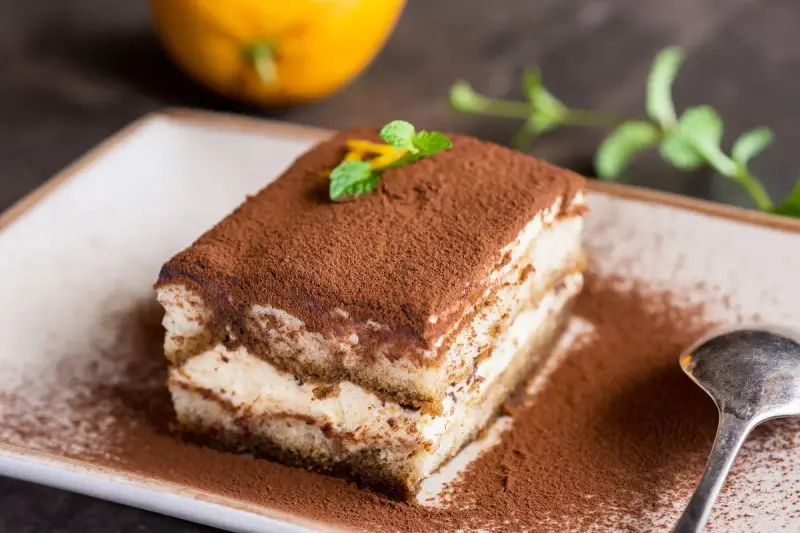 Pyszne tiramisu na biszkopcie, posypane kakao, z miętą i skórką pomarańczy. Obok łyżeczka i pomarańcza.